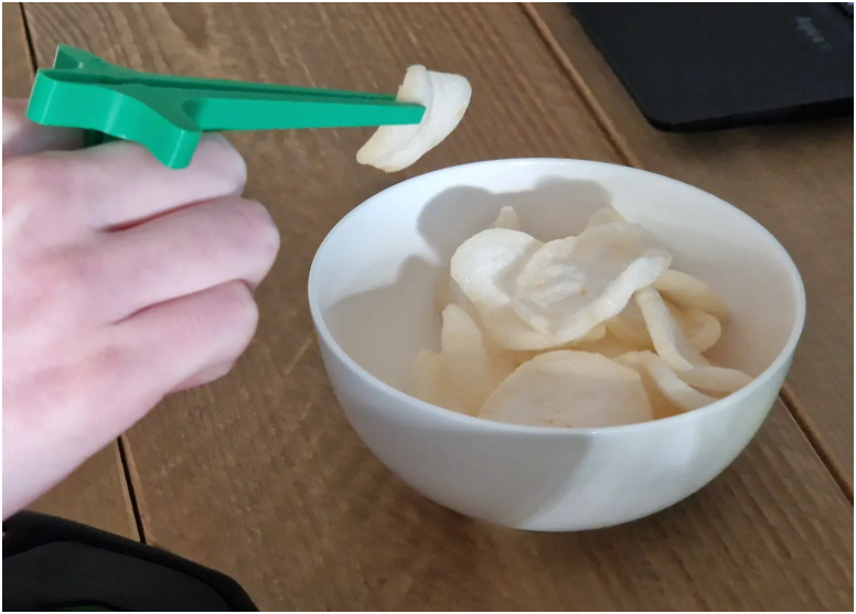 A hand holding a bright green plastic chip pick holder above a white bowl filled with potato chips on a wooden table.